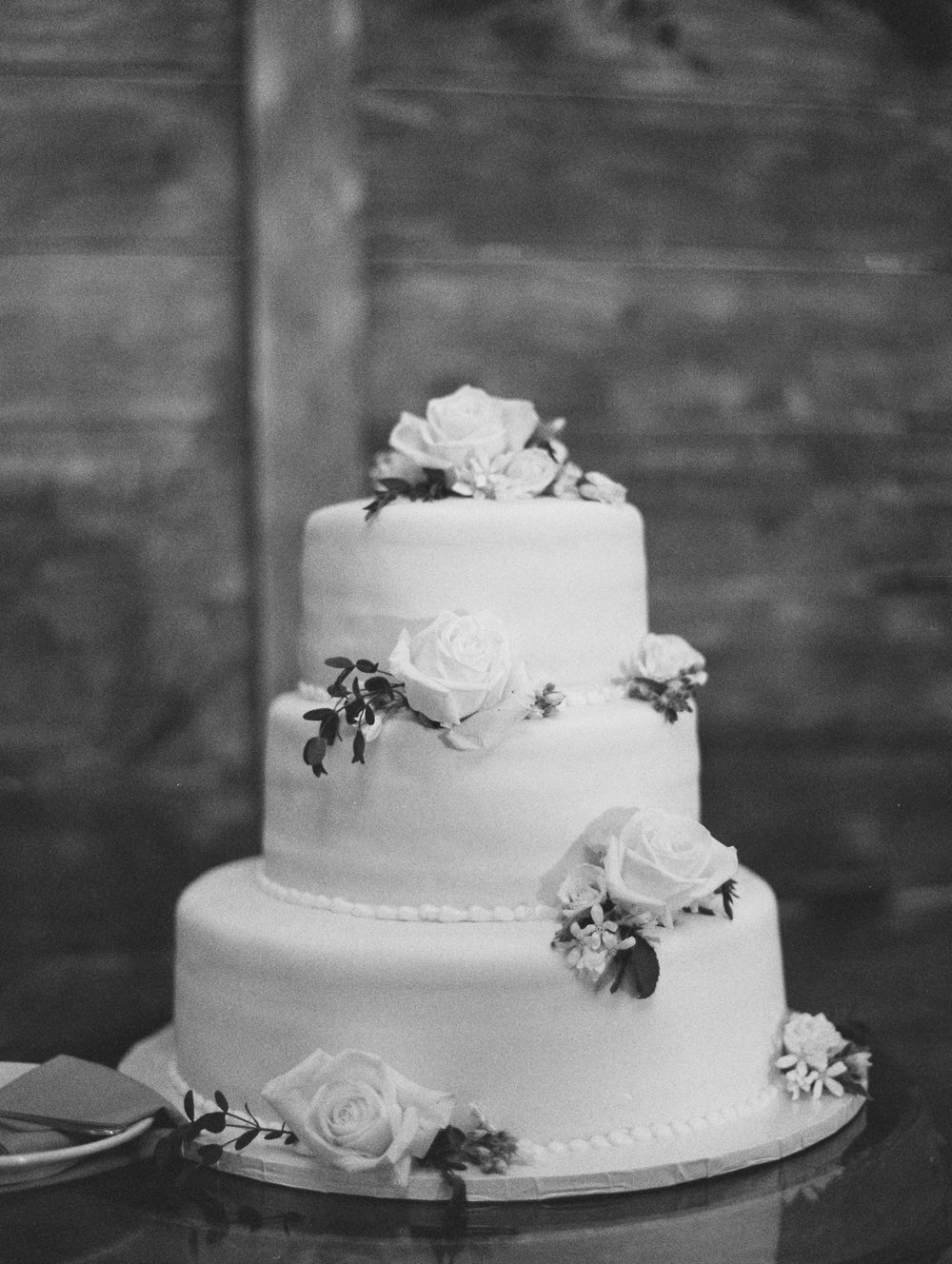 Classic three-tier white wedding cake adorned with white roses and greenery against rustic backdrop.