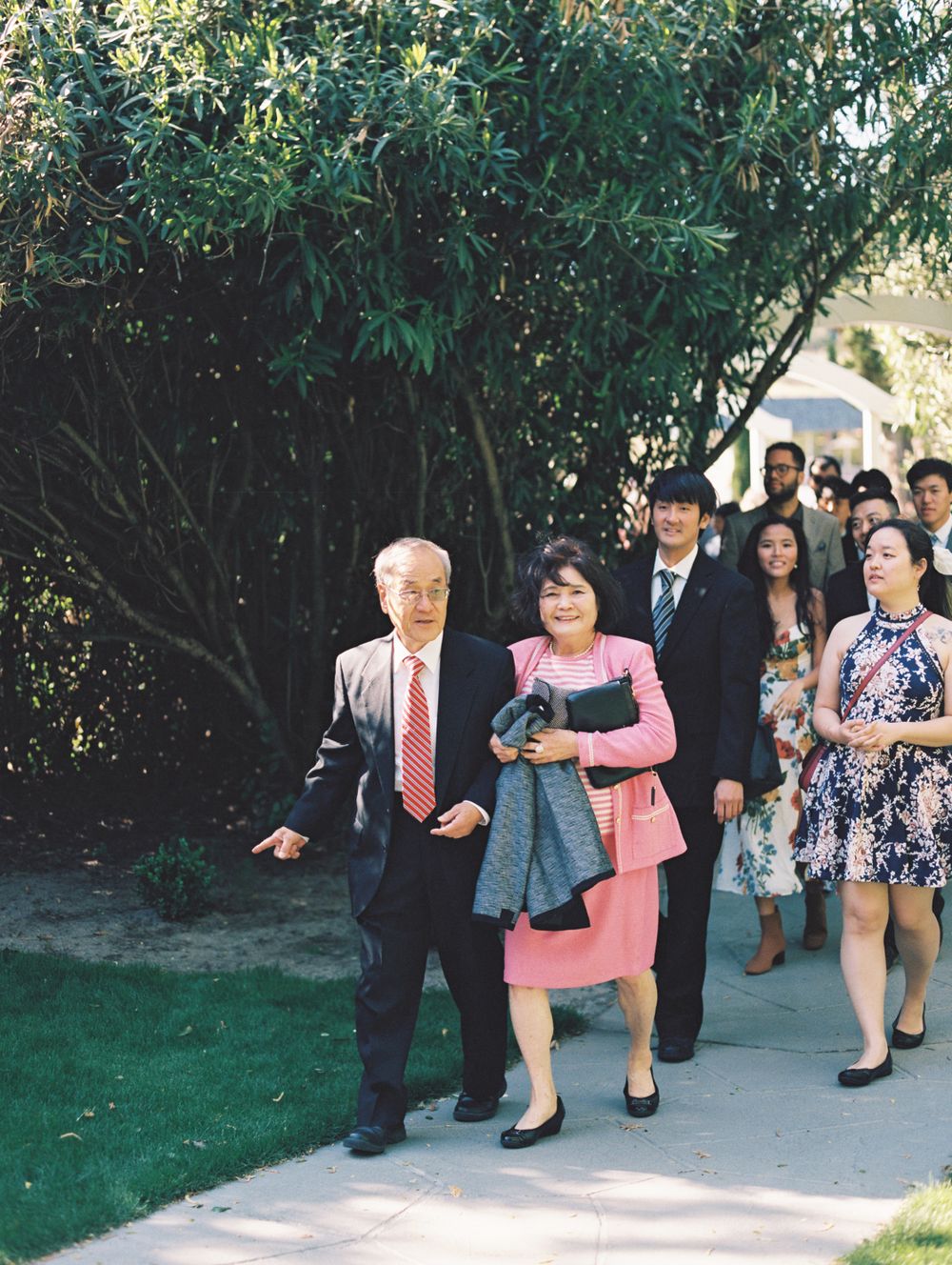 Formal wedding guests proceeding down an outdoor pathway surrounded by trees.