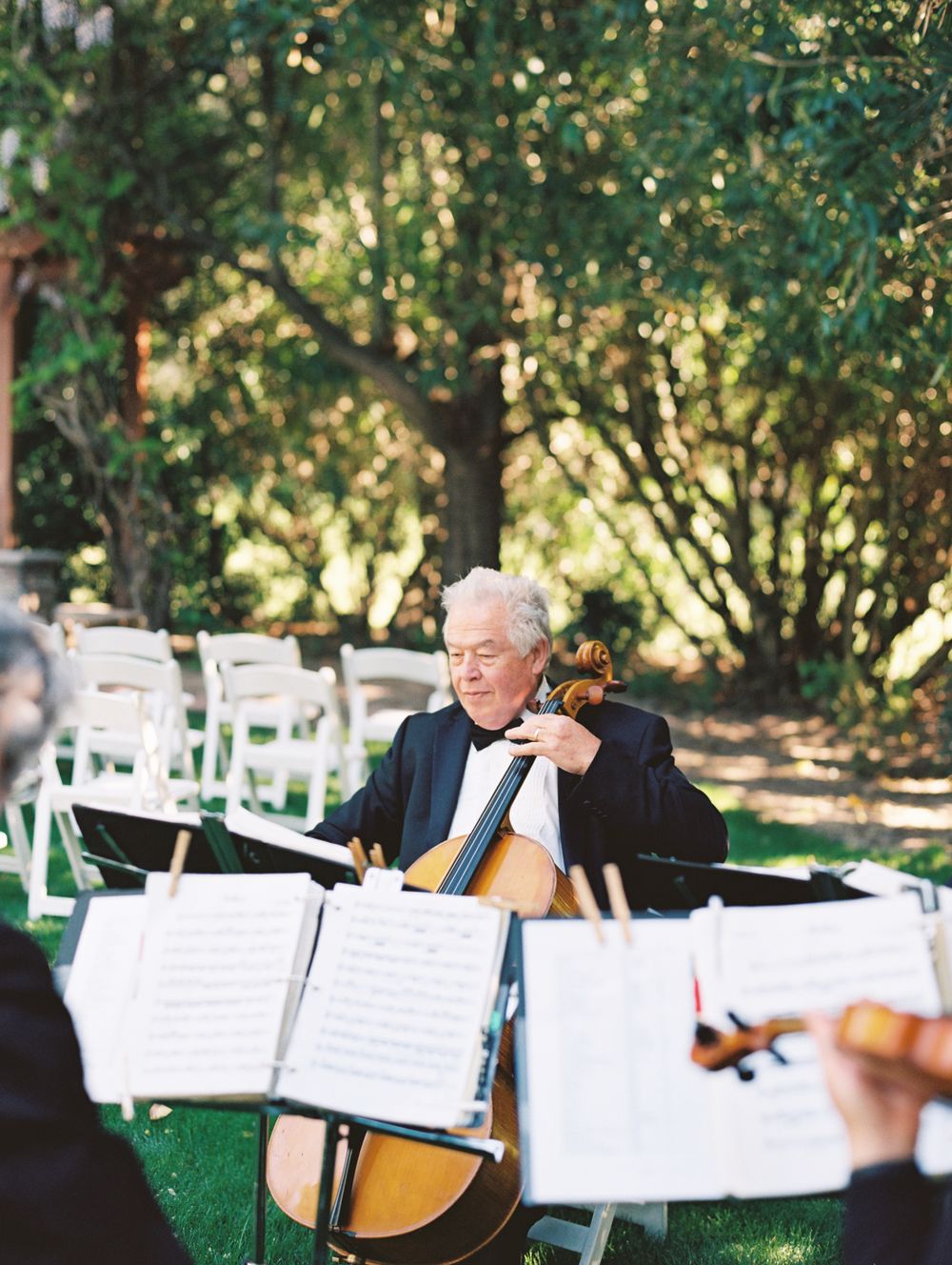 A cellist performs outdoors at a garden wedding ceremony with sheet music stands and white chairs.