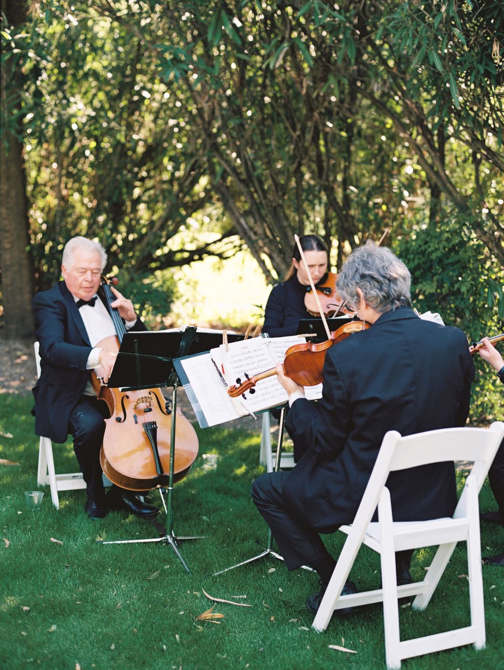 String trio performing classical music outdoors on white folding chairs.