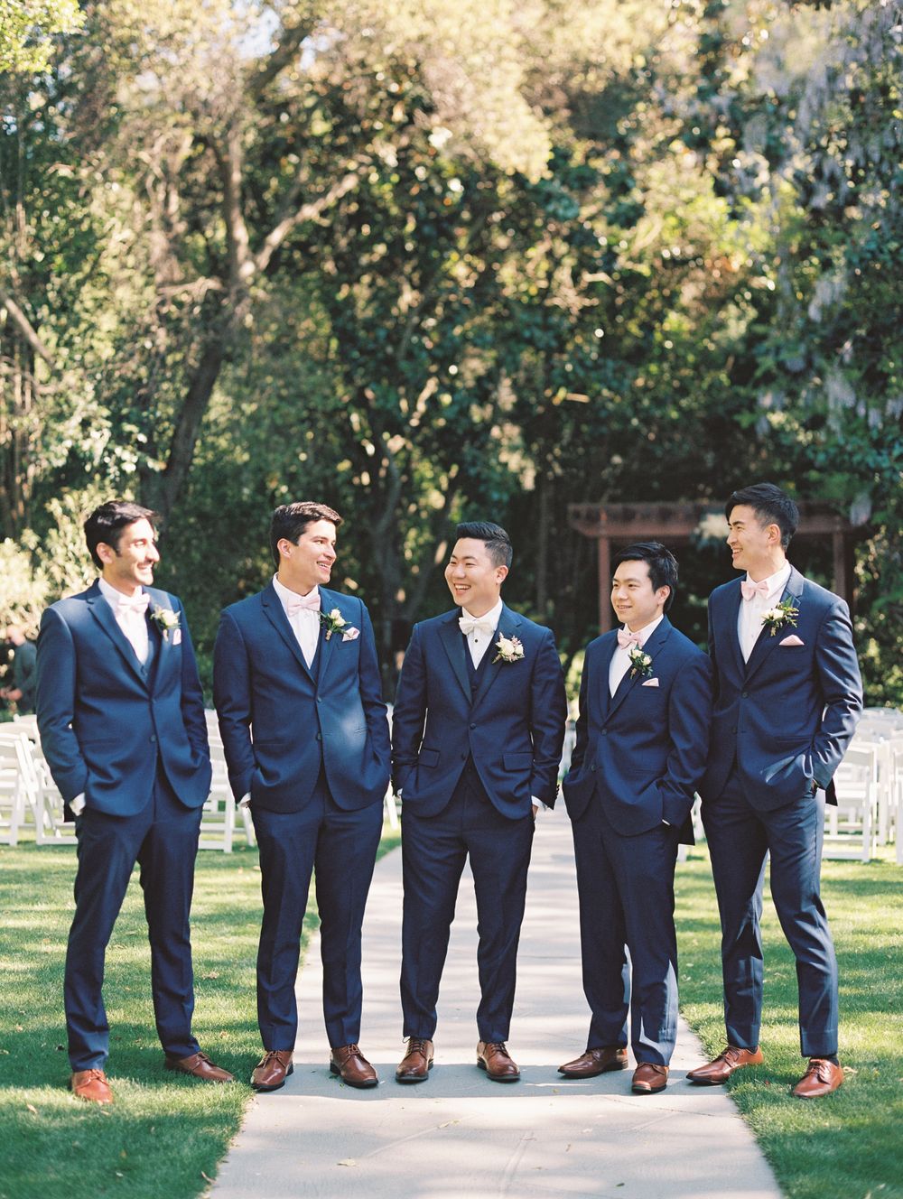 Five groomsmen in matching navy suits and brown shoes pose on a garden pathway.