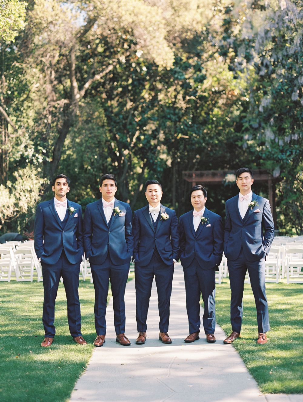 Groomsmen in navy blue suits standing together on garden pathway with white chairs in background.