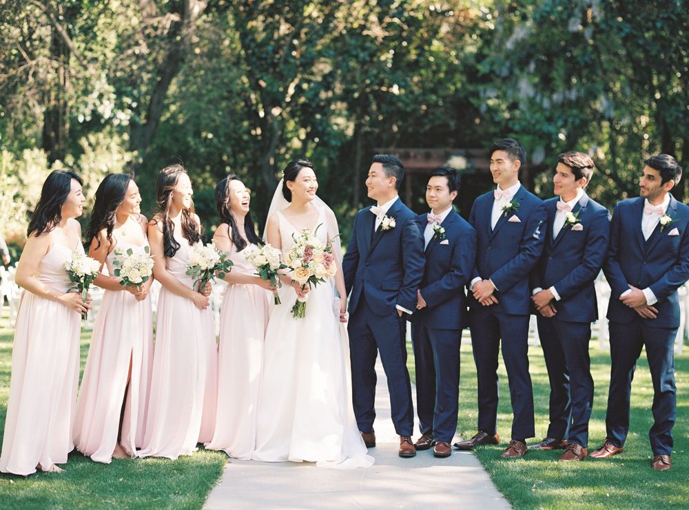 Wedding party in navy suits and blush pink dresses posing outdoors on garden pathway.