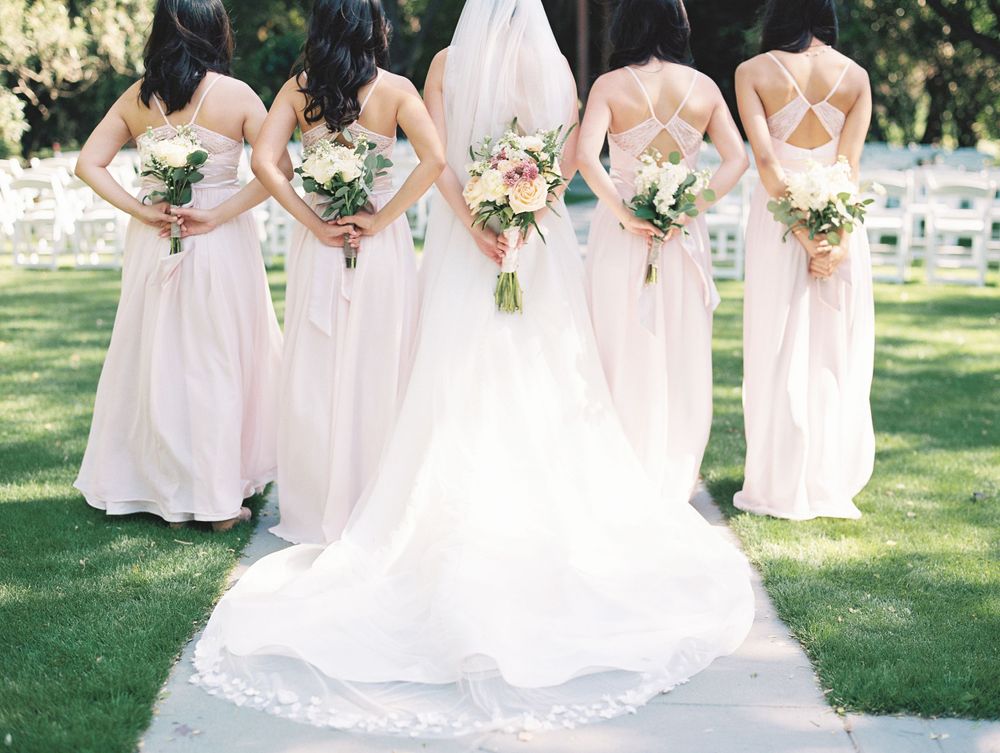 Bridesmaids in blush pink dresses holding bouquets behind bride with flowing white train.