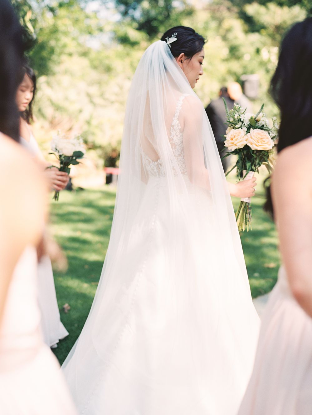 Bride in white lace gown with long veil surrounded by bridesmaids holding bouquets.