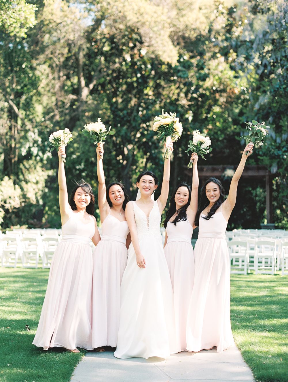 Bridal party in pink dresses raising white bouquets joyfully on garden pathway.