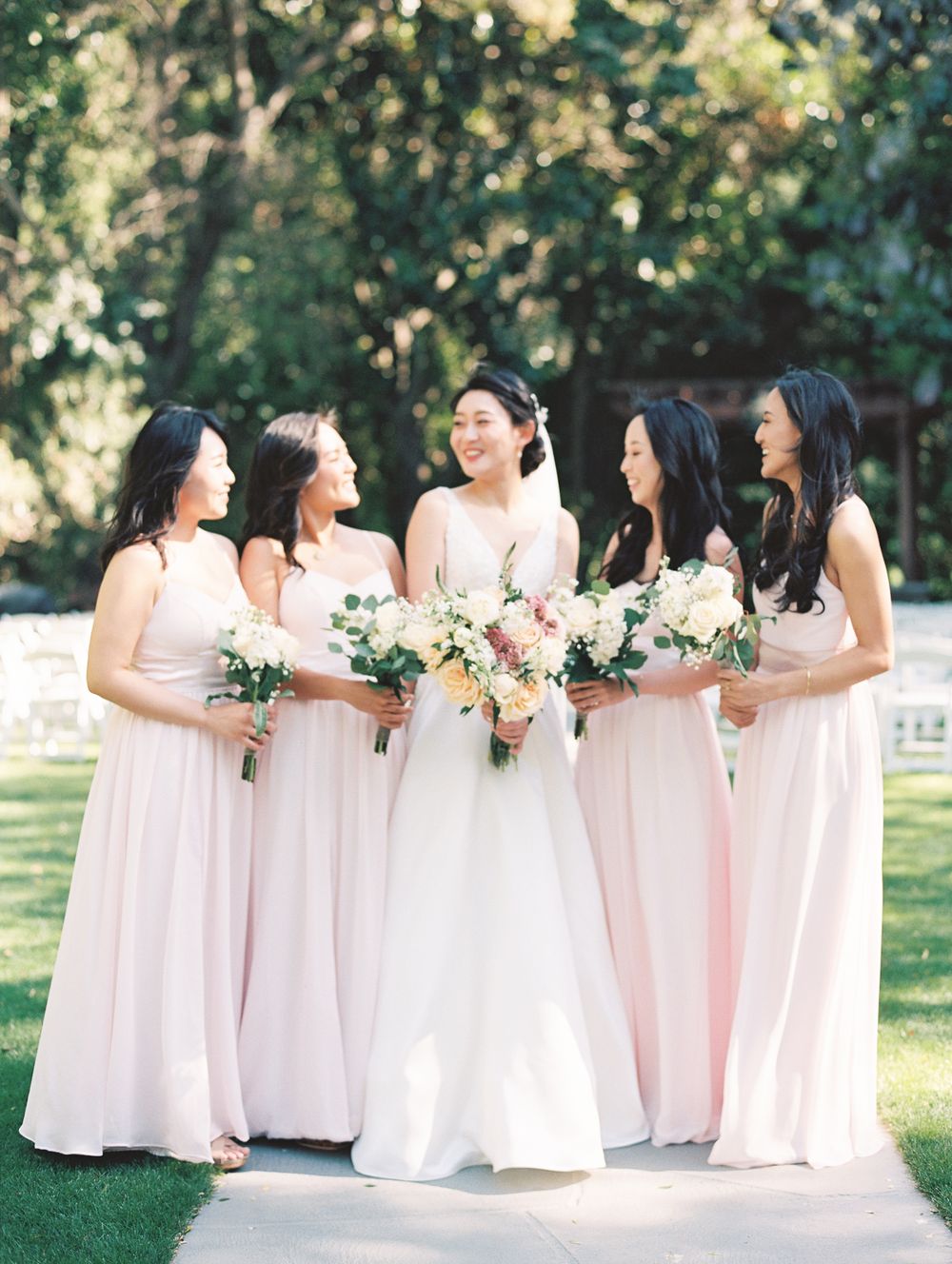 Bride in white gown with bridesmaids in blush pink dresses holding floral bouquets outdoors.