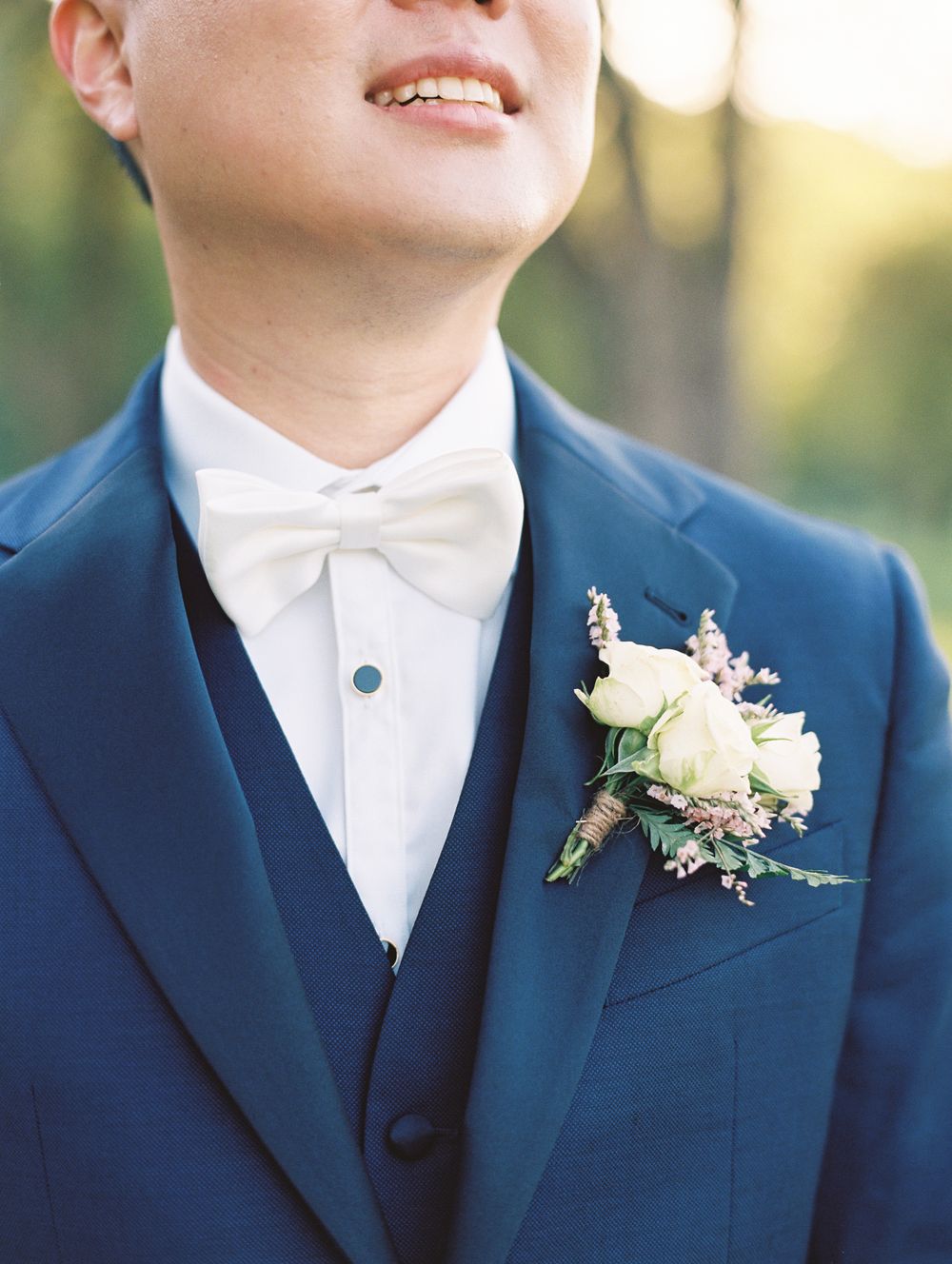 Wedding attire detail showing navy suit with white bowtie and delicate floral boutonniere in natural lighting.