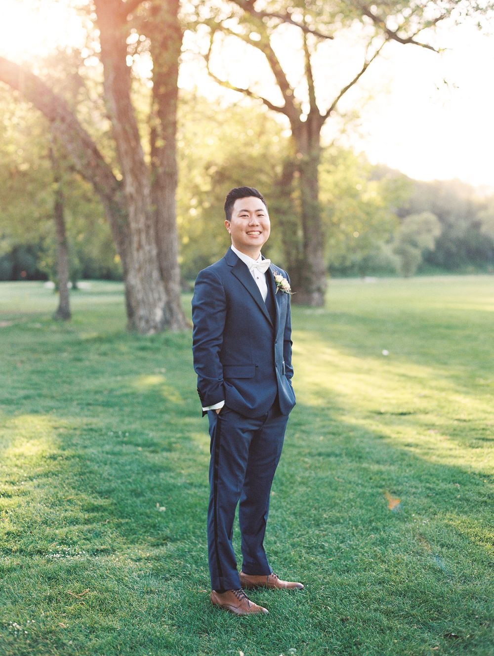 A formal wedding portrait outdoors with natural sunlight filtering through trees in the background.