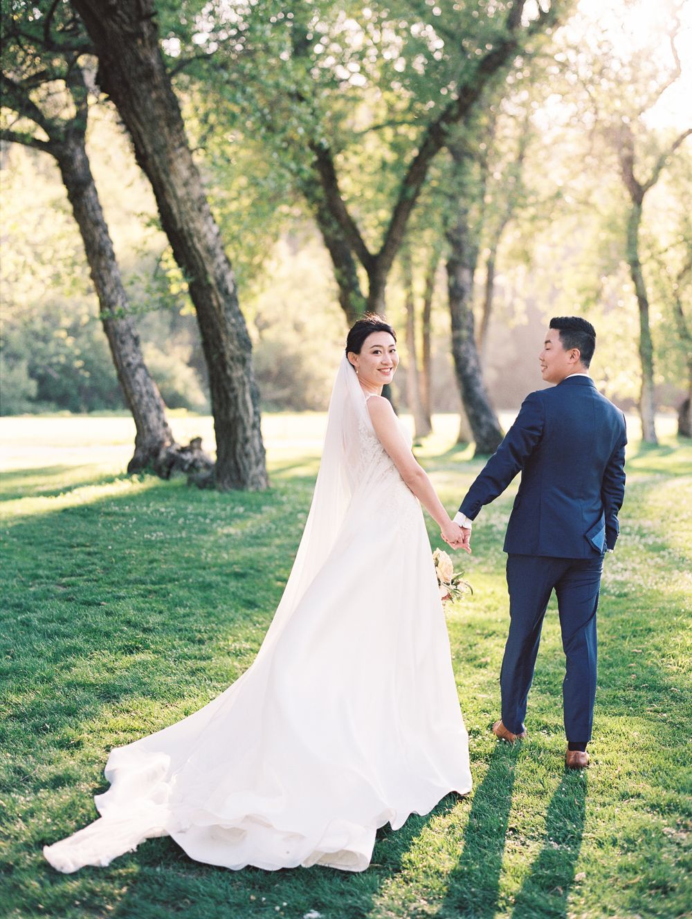 A wedding couple walks on grass beneath tall trees in a park while holding hands.