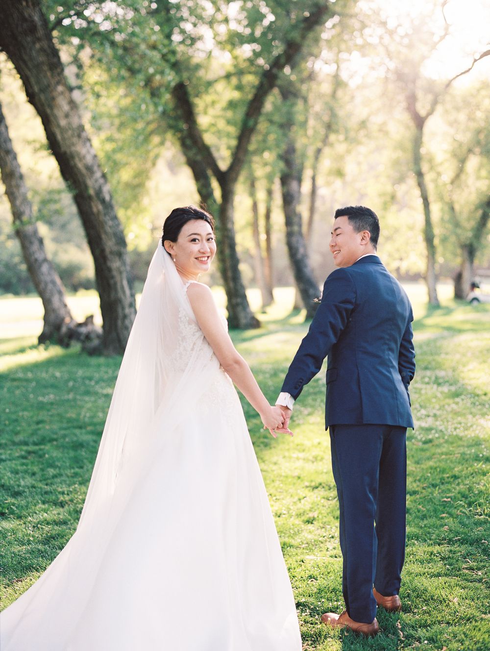A bride in a flowing white gown and groom in navy suit pose together in a sunlit outdoor setting.
