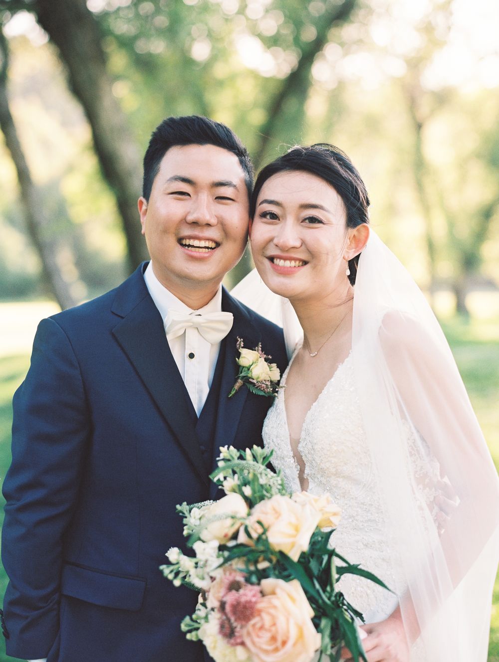 A bride in white dress smiles while holding a peach and white rose bouquet outdoors.