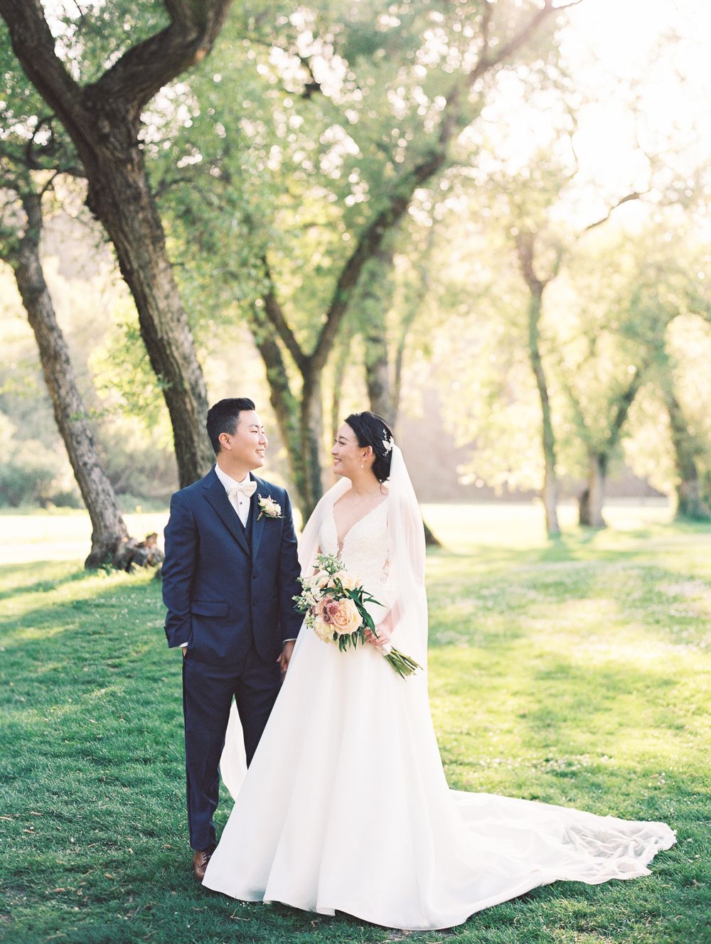 A wedding couple poses together on green grass with trees in soft natural lighting.