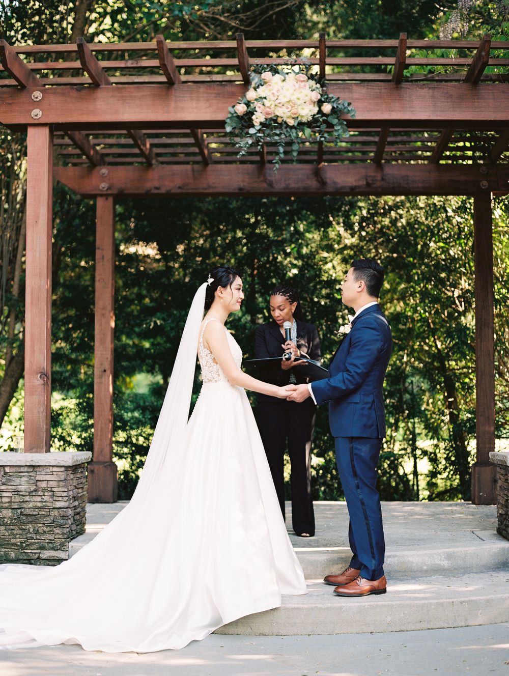 An outdoor wedding ceremony under a wooden pergola decorated with white floral arrangements.