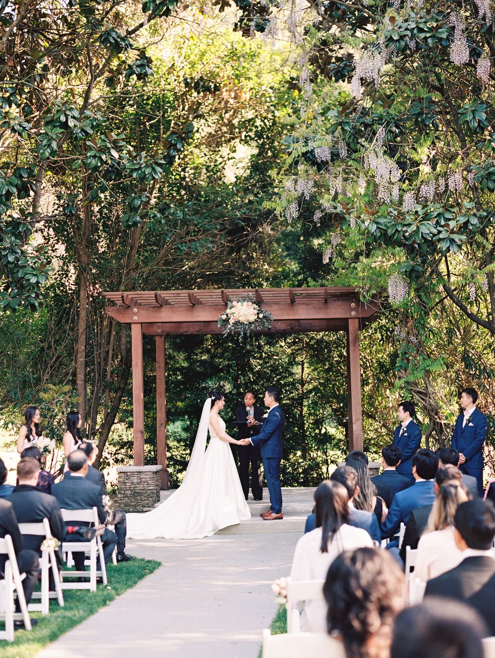 An outdoor wedding ceremony under a rustic wooden pergola surrounded by lush greenery.
