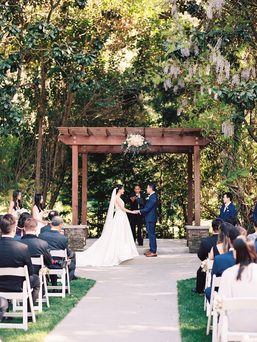 Guests seated in white chairs watch a wedding ceremony under a wooden pergola in a garden setting.