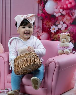 A pink-themed baby photo session setup with a wicker basket and teddy bear on a plush armchair.