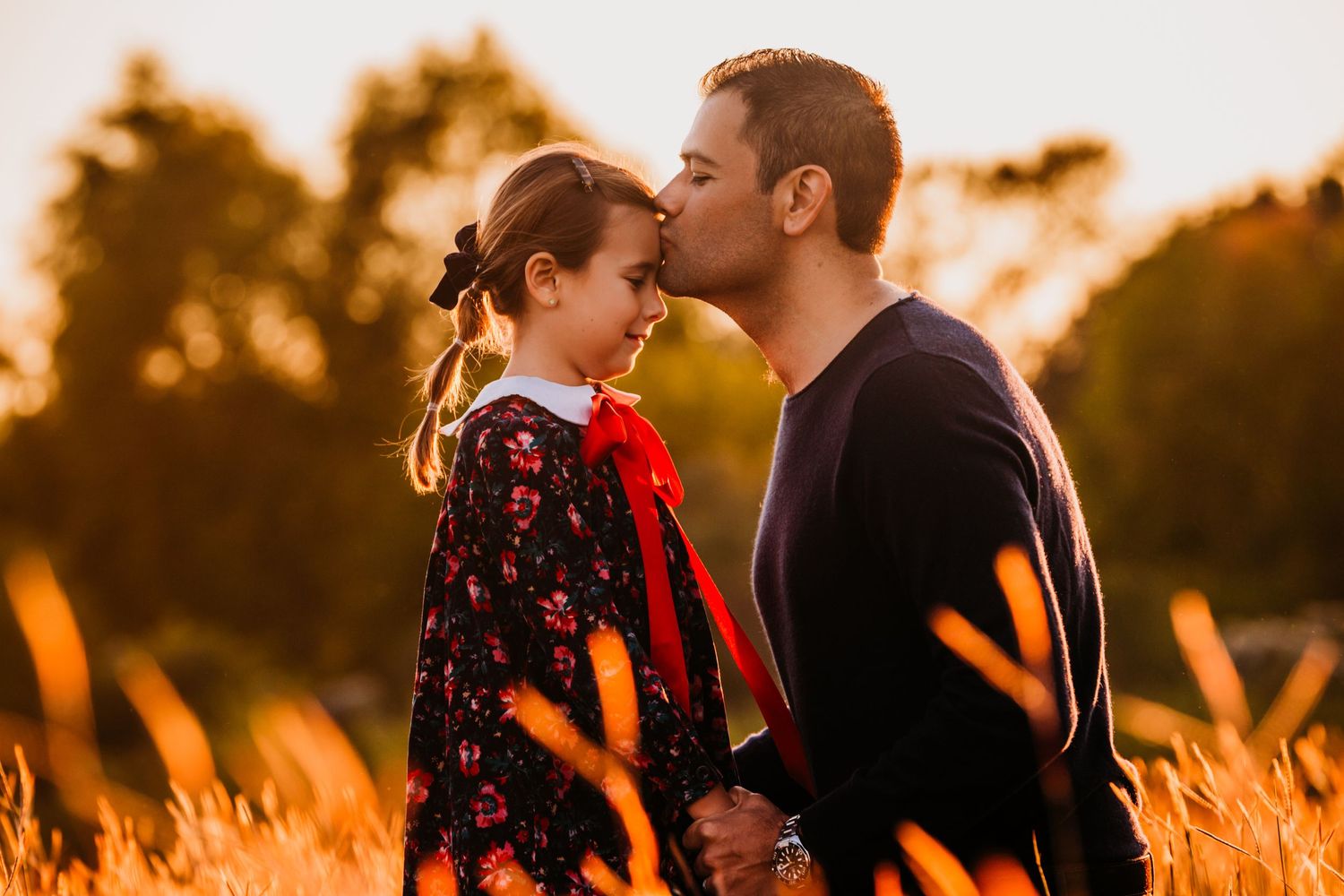 A dad and daughter shares a tender moment in a golden wheat field at sunset while wearing casual attire.