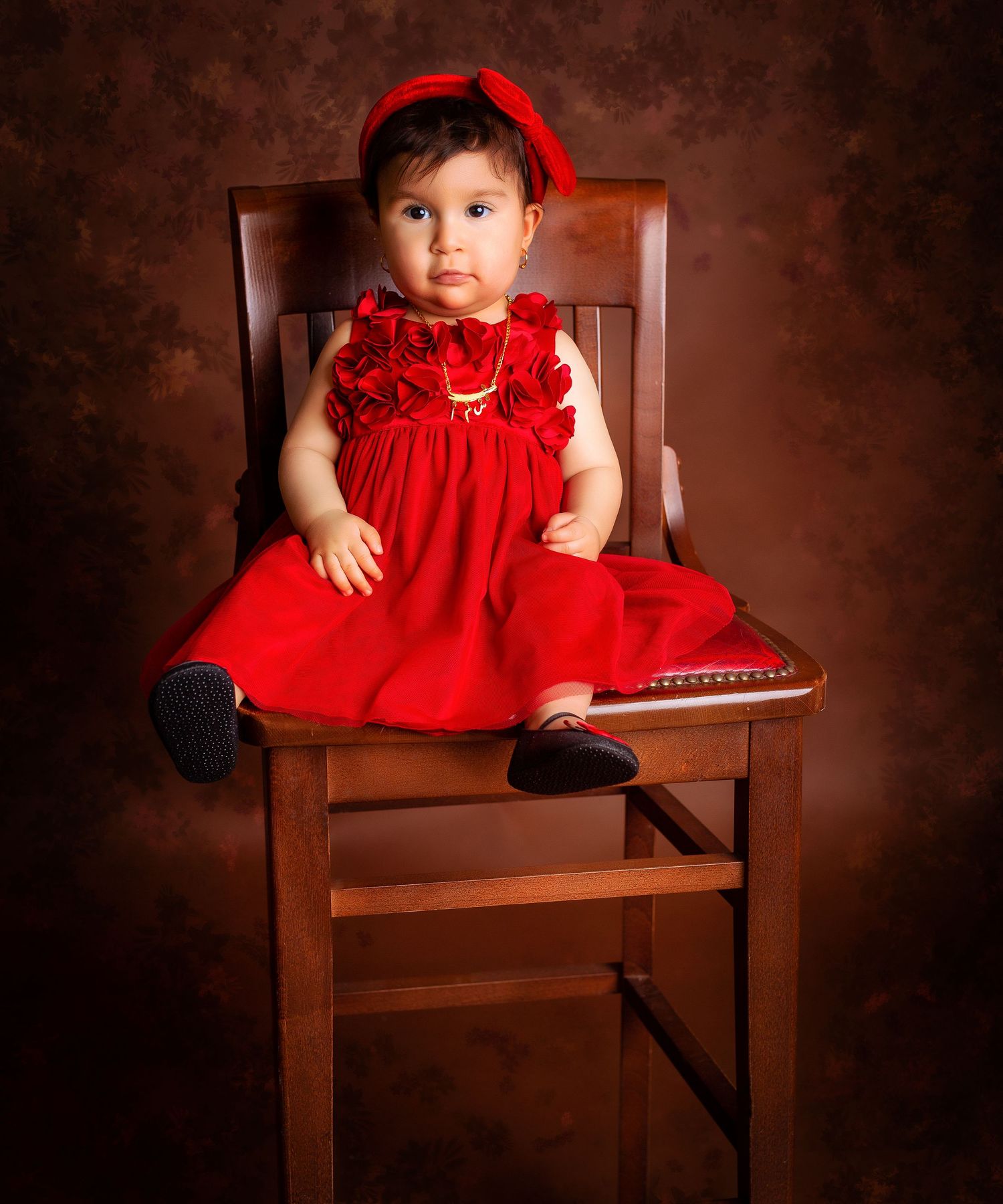 A little girl in flowery red dress is sitting on a high chair and looking into the camera with a brown background, Great toddler portrait