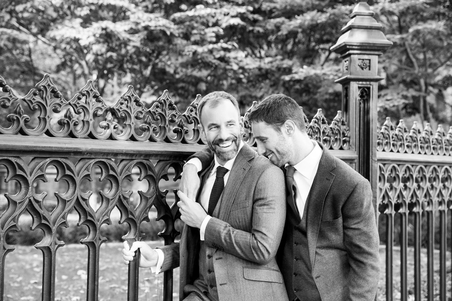 Two men in suits share a lighthearted moment by an ornate iron fence.