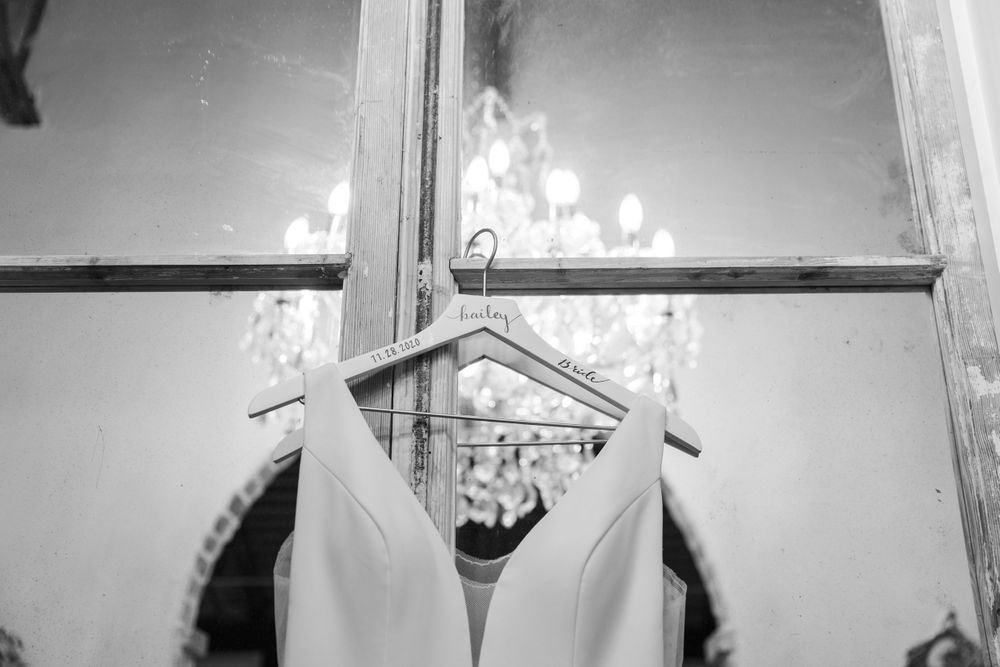 A white wedding dress hangs in front of a window with a chandelier reflection.