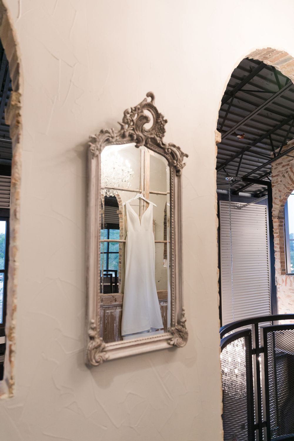 A white wedding dress hangs in an ornate vintage mirror against a light-colored wall.