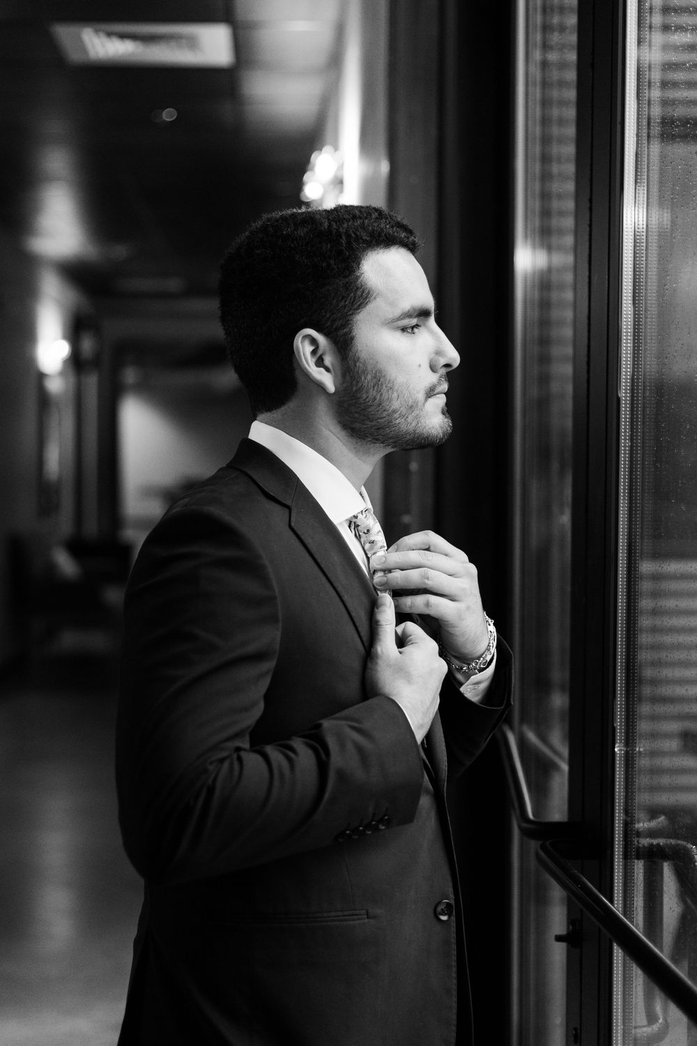Black and white portrait of a well-dressed person adjusting their tie near a window.