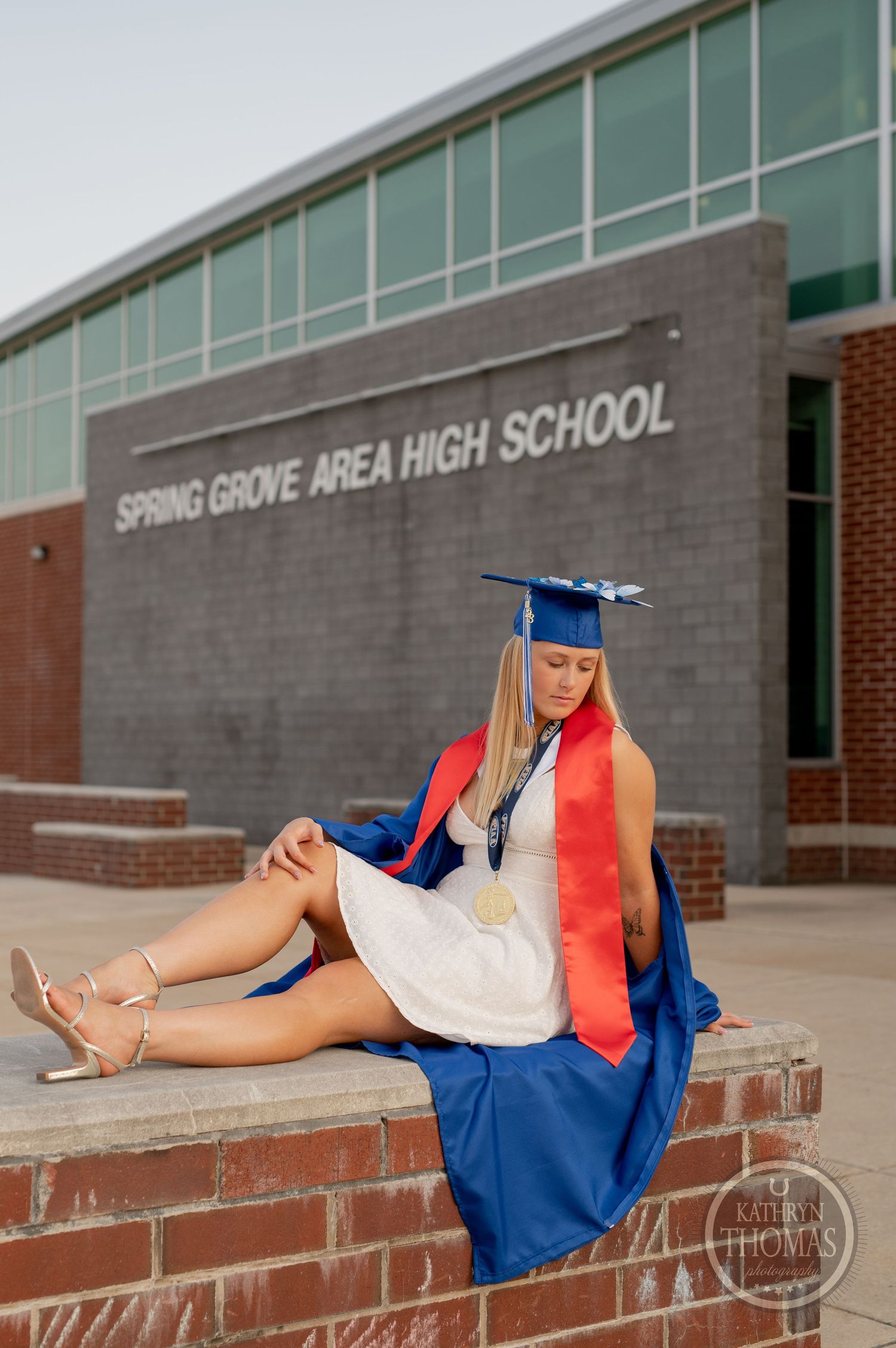 Graduate in white dress with blue gown and red stole poses on brick ledge at high school.
