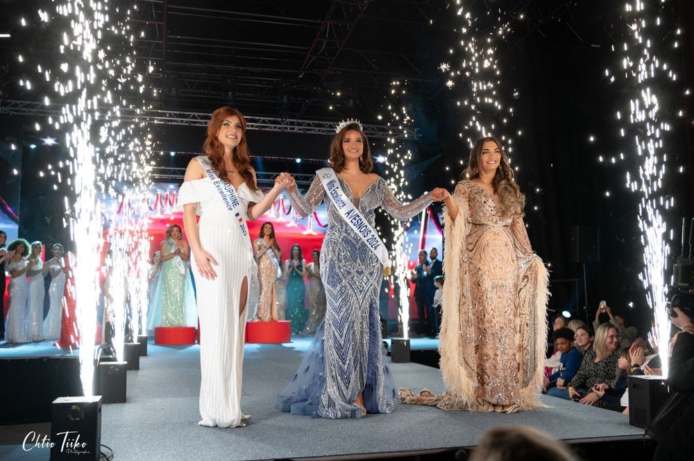 Three pageant finalists in evening gowns stand on stage with fireworks display.