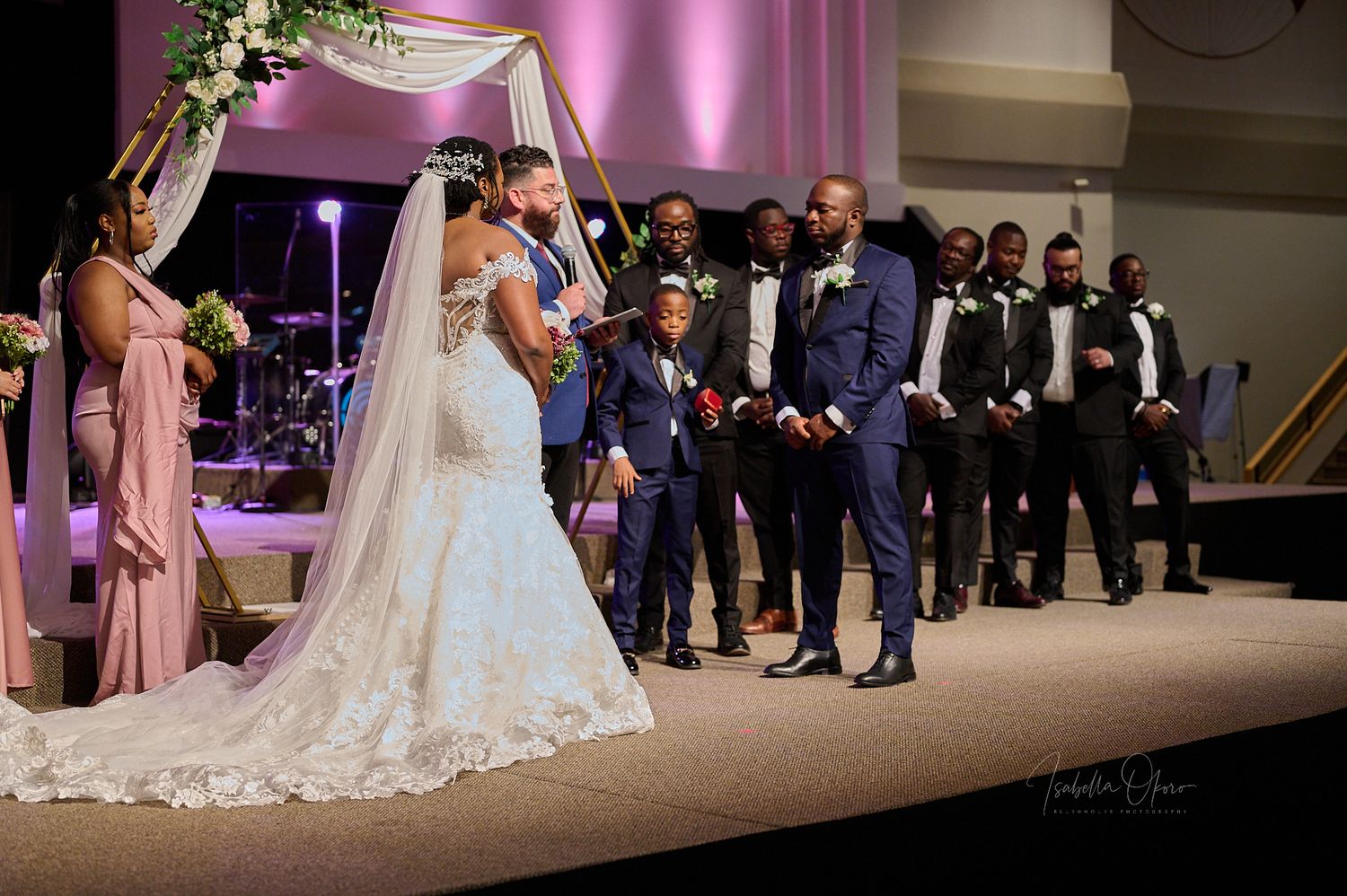 Wedding ceremony moment with bridal party and groomsmen lined up on stage under decorative arch.