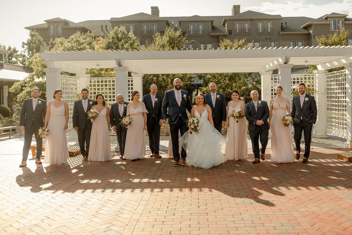 Wedding party arranged in formal line under white pergola with greenery.