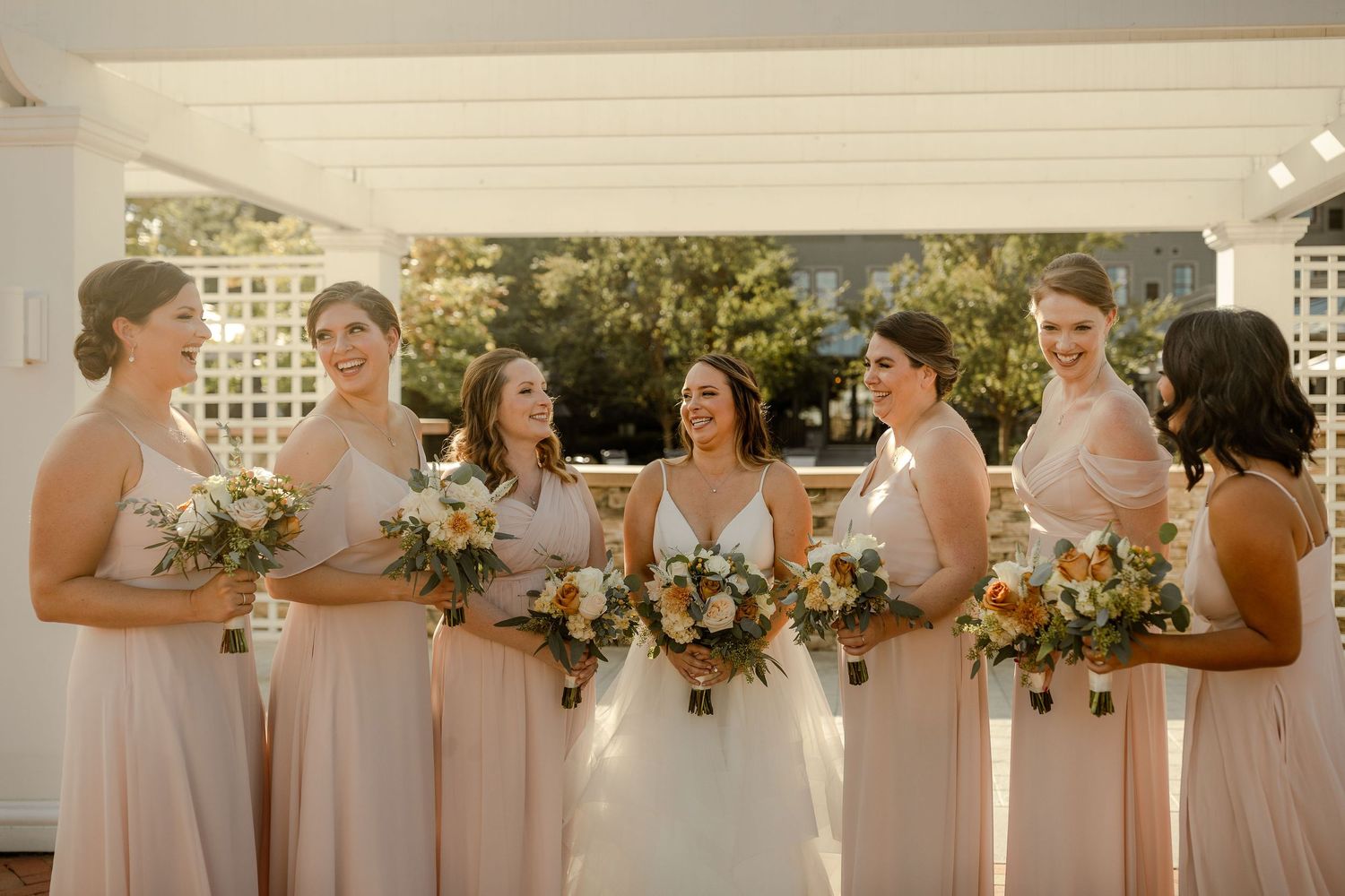 Bridesmaids in blush pink dresses holding bouquets under white pergola.