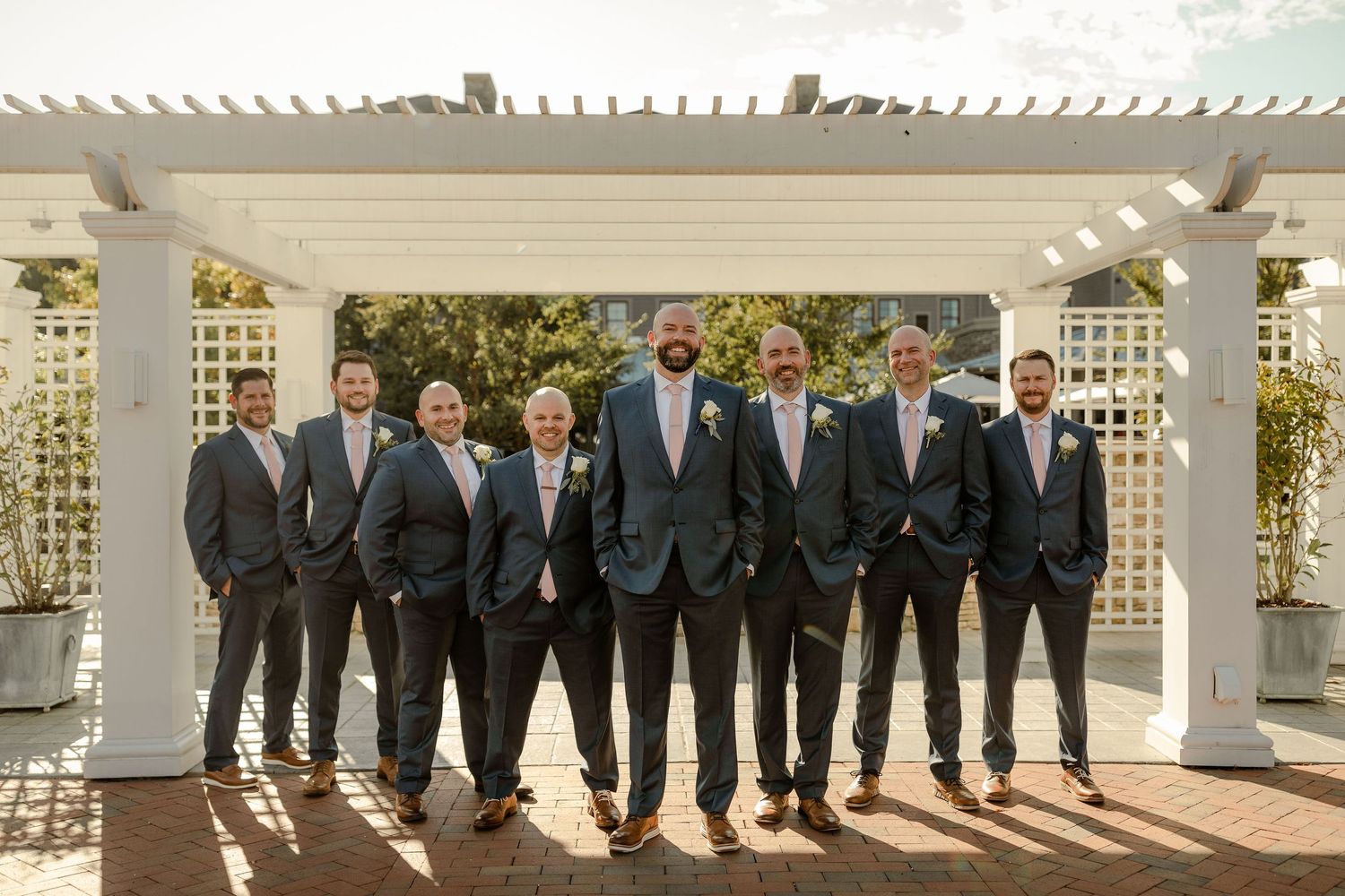 Groomsmen in dark suits lined up under white pergola for formal wedding photo.
