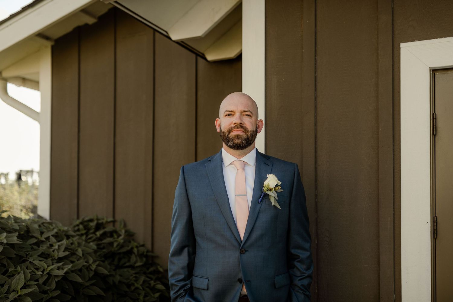 Groomsmen in matching navy suits posing against rustic brown wall at outdoor venue.