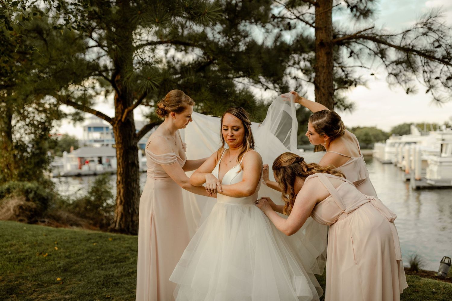 Bridesmaids in blush pink dresses gathered around bride in flowing white gown under pine trees.