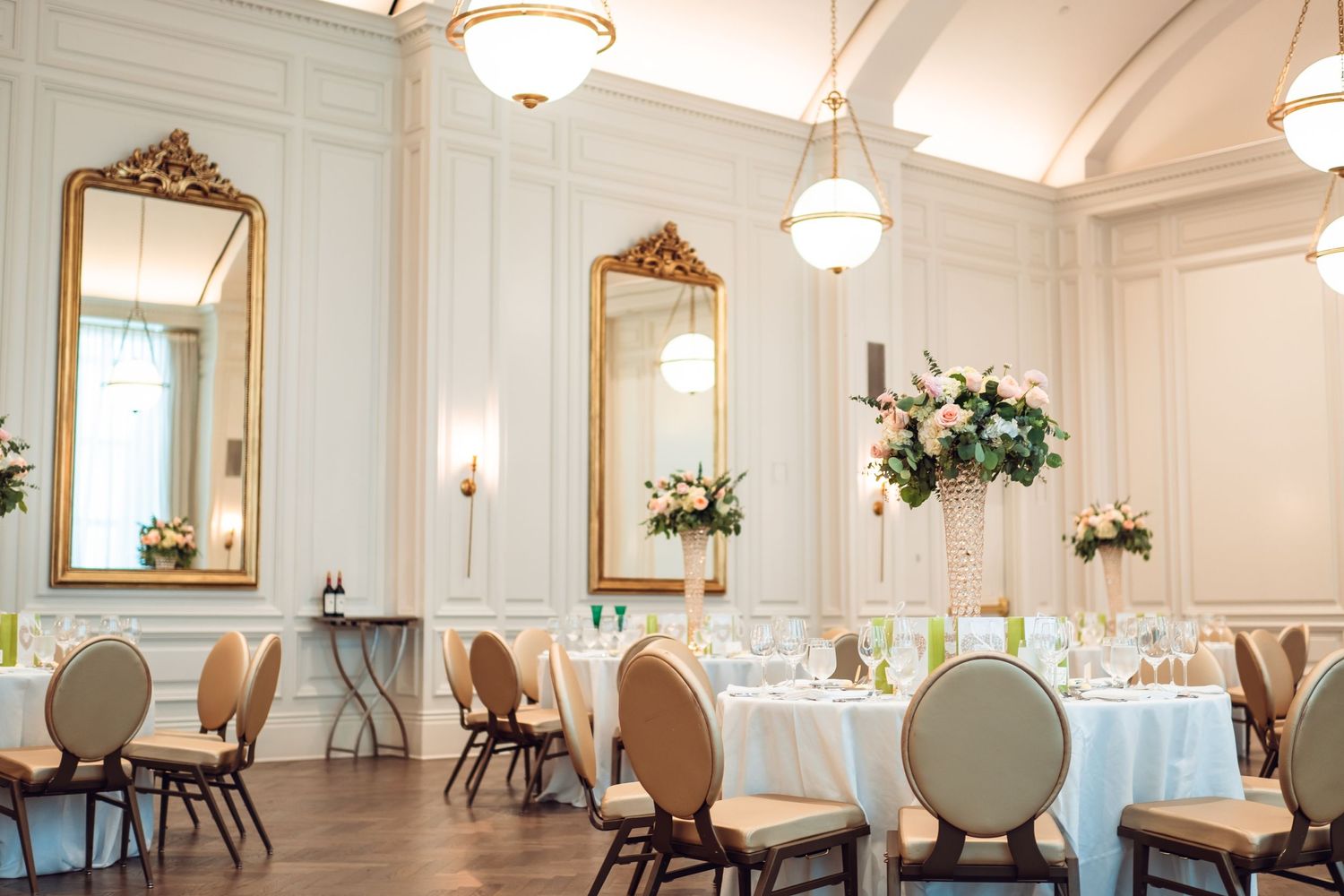Elegant white ballroom with ornate mirrors, chandeliers and round tables set for a formal event.