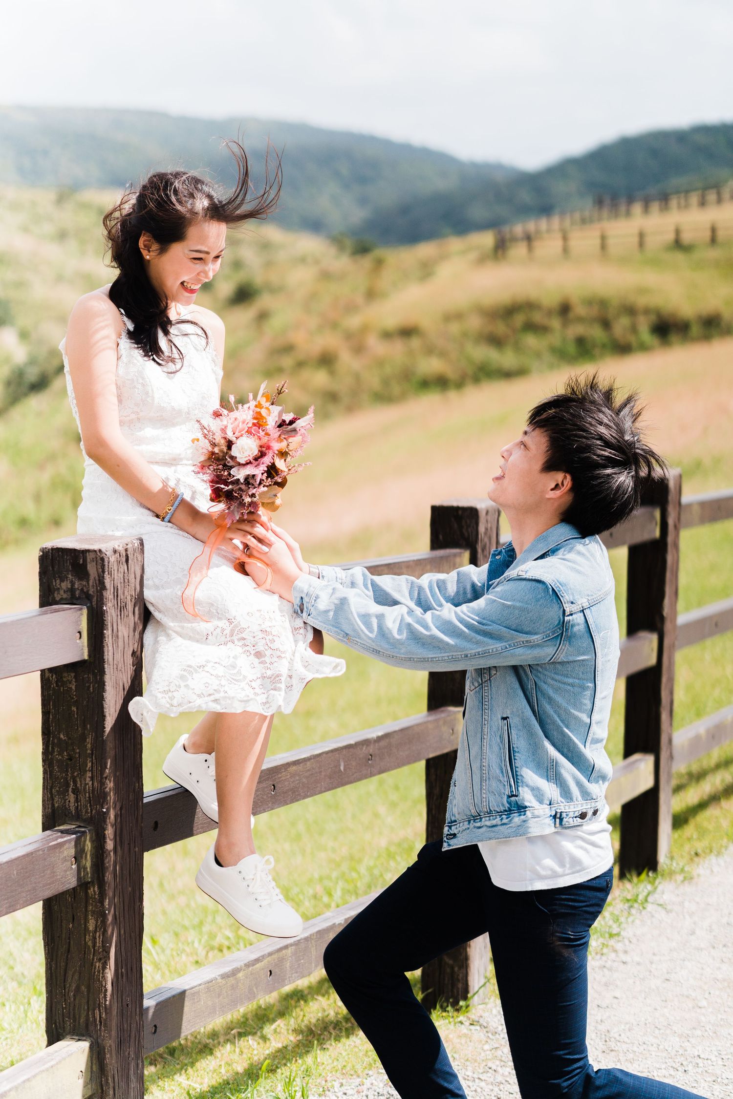 Couple sharing romantic moment on wooden fence with flowers and mountains in background.