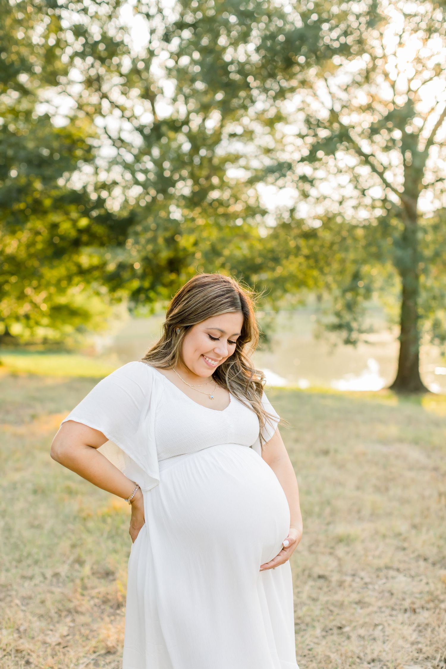 Side view of pregnant woman in white dress cradling baby bump in outdoor nature setting.