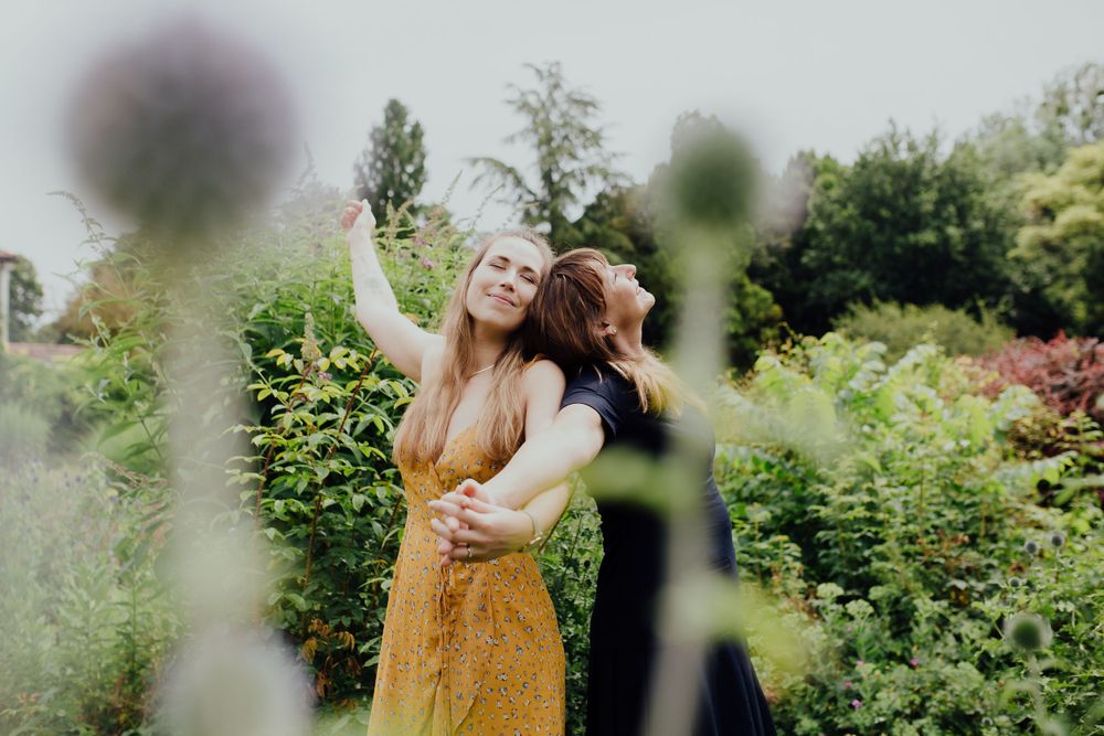 Eine Person in braunen Latzhosen tanzt fröhlich in einem üppigen Sommergarten mit Blumen und Grünpflanzen.