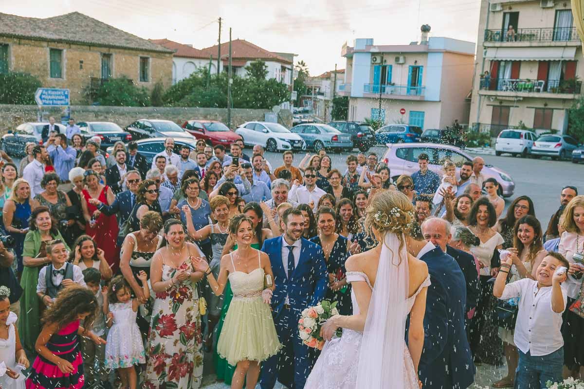 A crowd of wedding guests smiling and gathering together in a parking lot during a joyful celebration by GeorgeNafpliotis.com