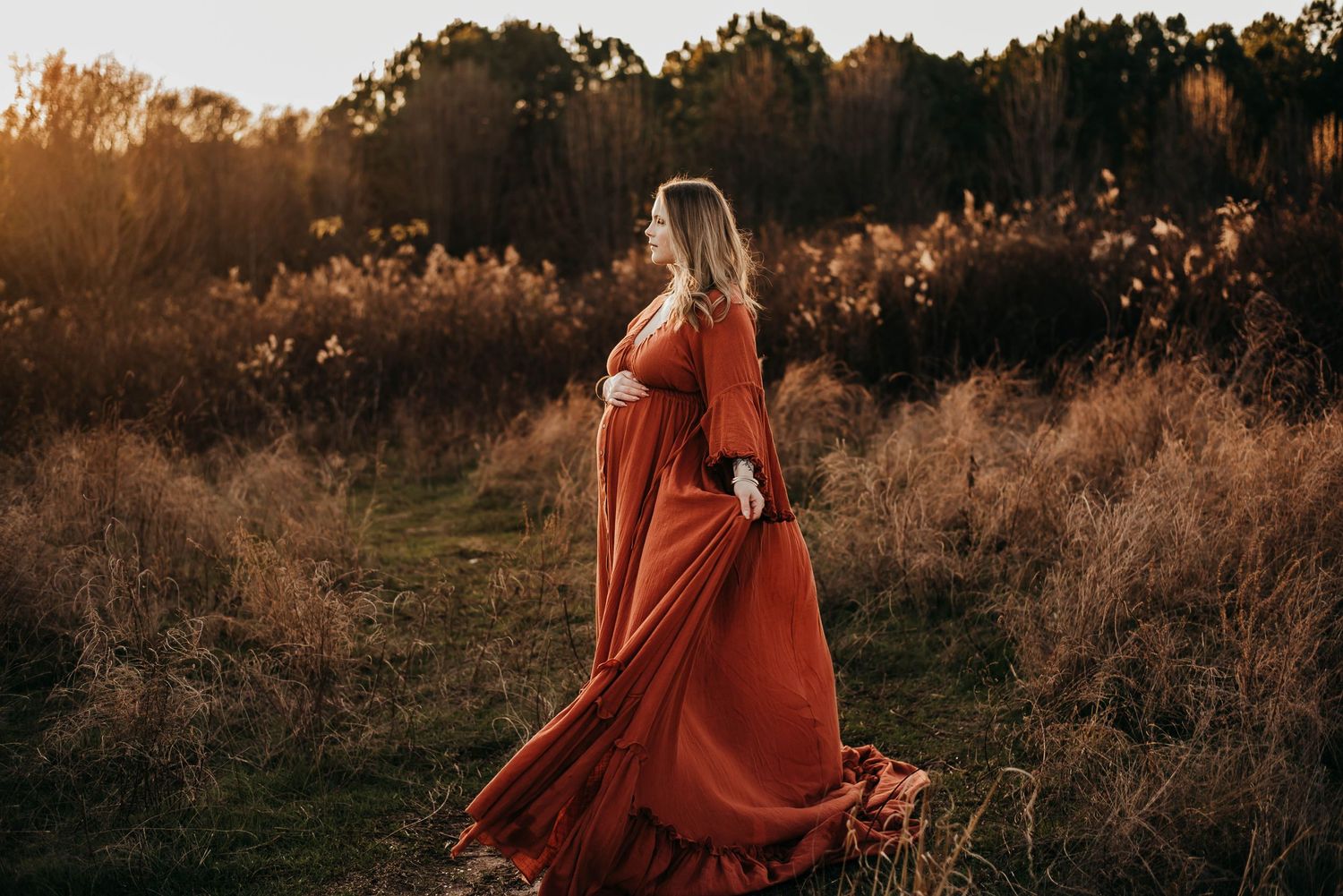 Pregnant woman in flowing rust-red gown standing in golden autumn field at sunset.