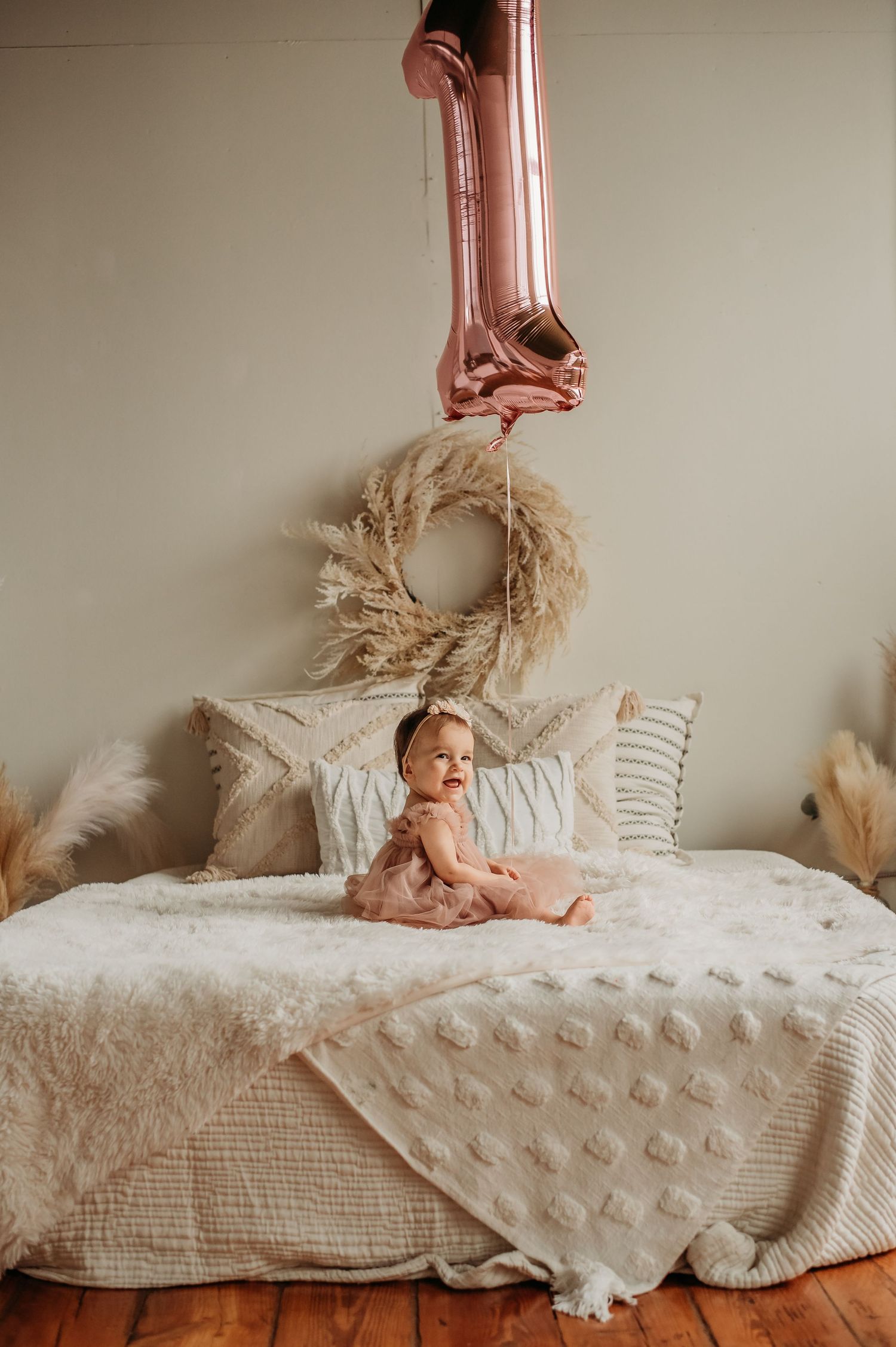Baby girl in pink tutu sits on bed with number one balloon for first birthday celebration.