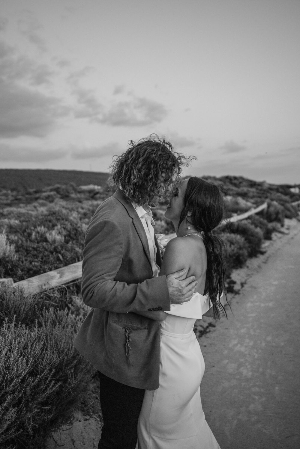 Romantic black and white photo of a couple embracing on a coastal path at sunset.
