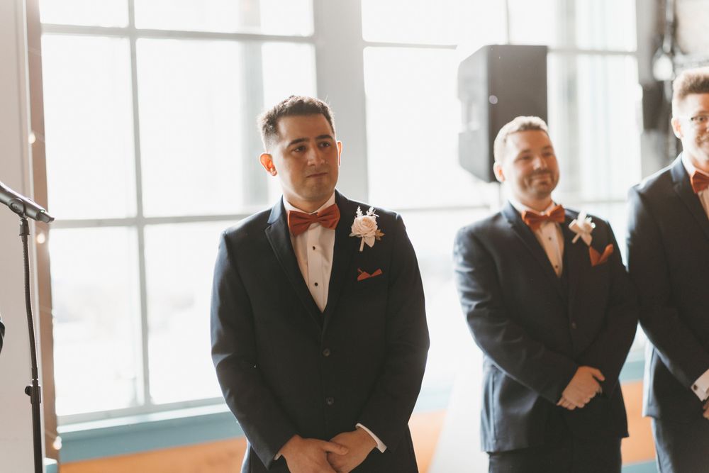 Groom and groomsmen in navy suits with orange bow ties standing at altar during wedding ceremony.