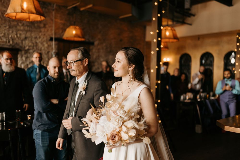Bride in white gown holding neutral bouquet walking down aisle with escort in rustic wedding venue.