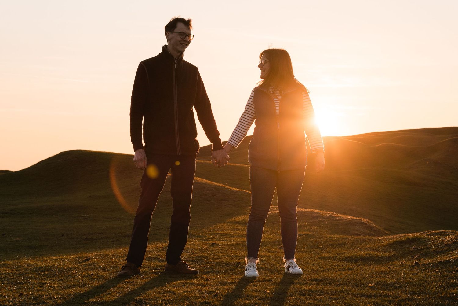 A young couple walking towards the camera over hills, at golden hour. The sun shining behind the. They are looking at each other and smiling