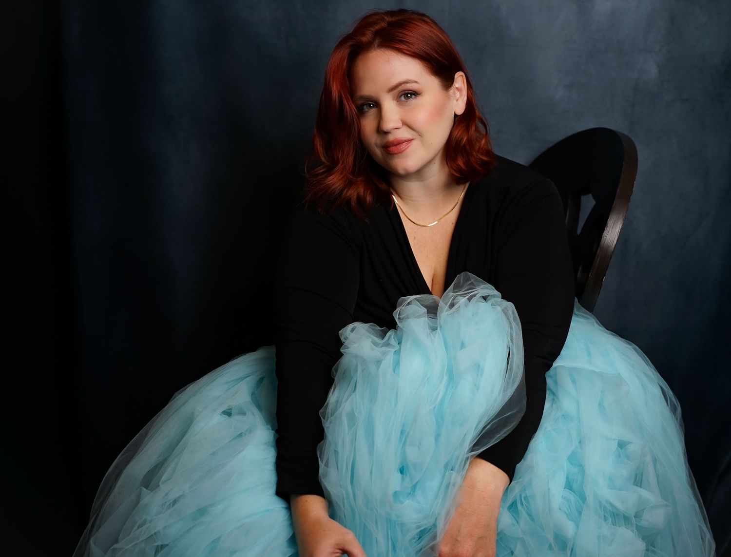 Woman in black jacket and blue tulle skirt poses elegantly against dark backdrop.