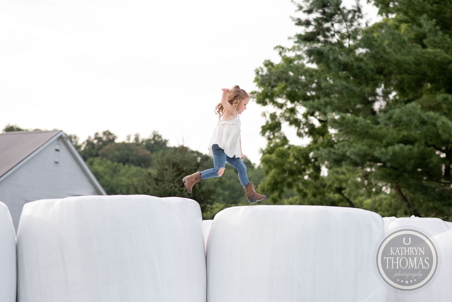 Child jumps across large white hay bales at sunset on a farm.