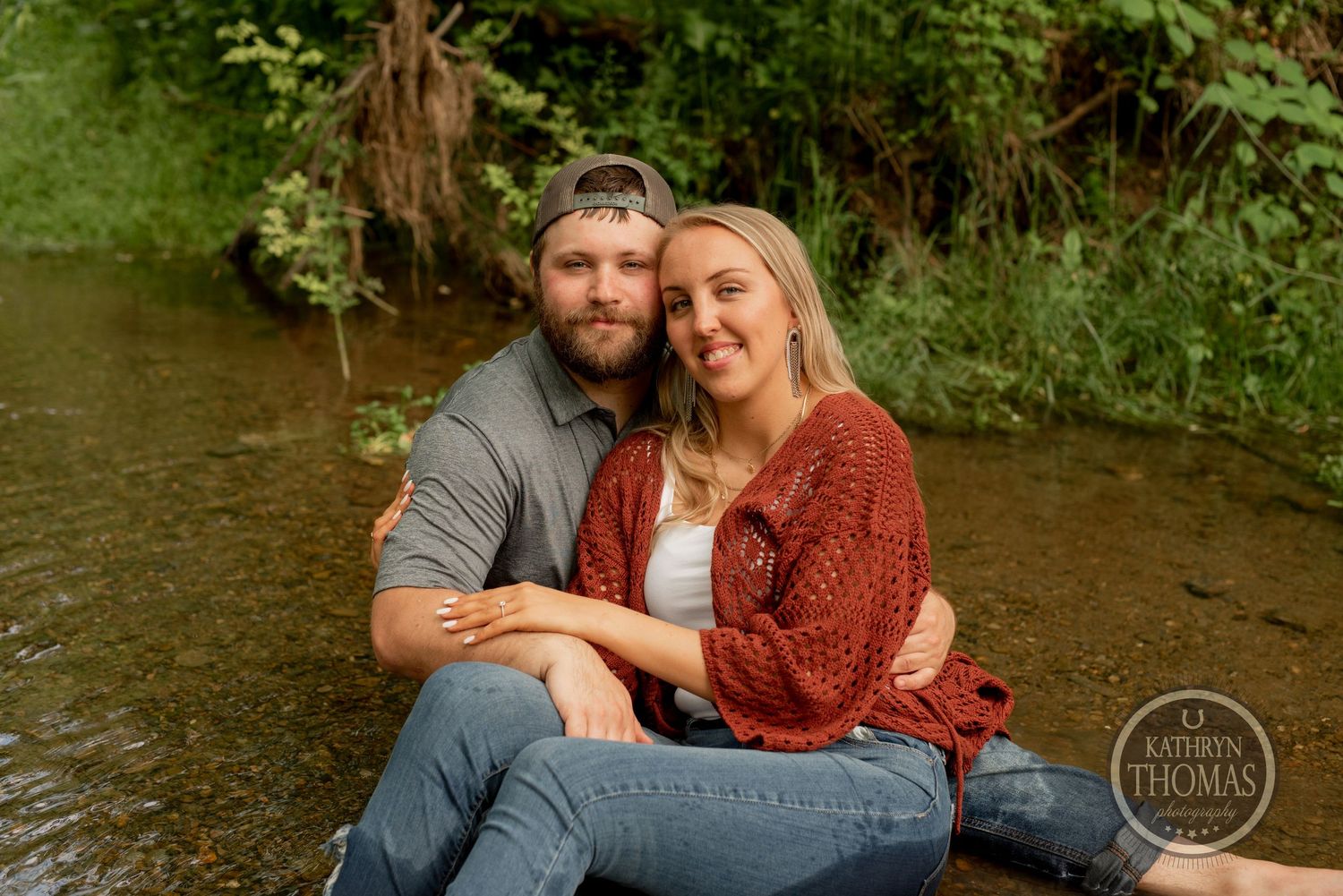 A couple sits together on rocks in a creek while surrounded by lush forest scenery.