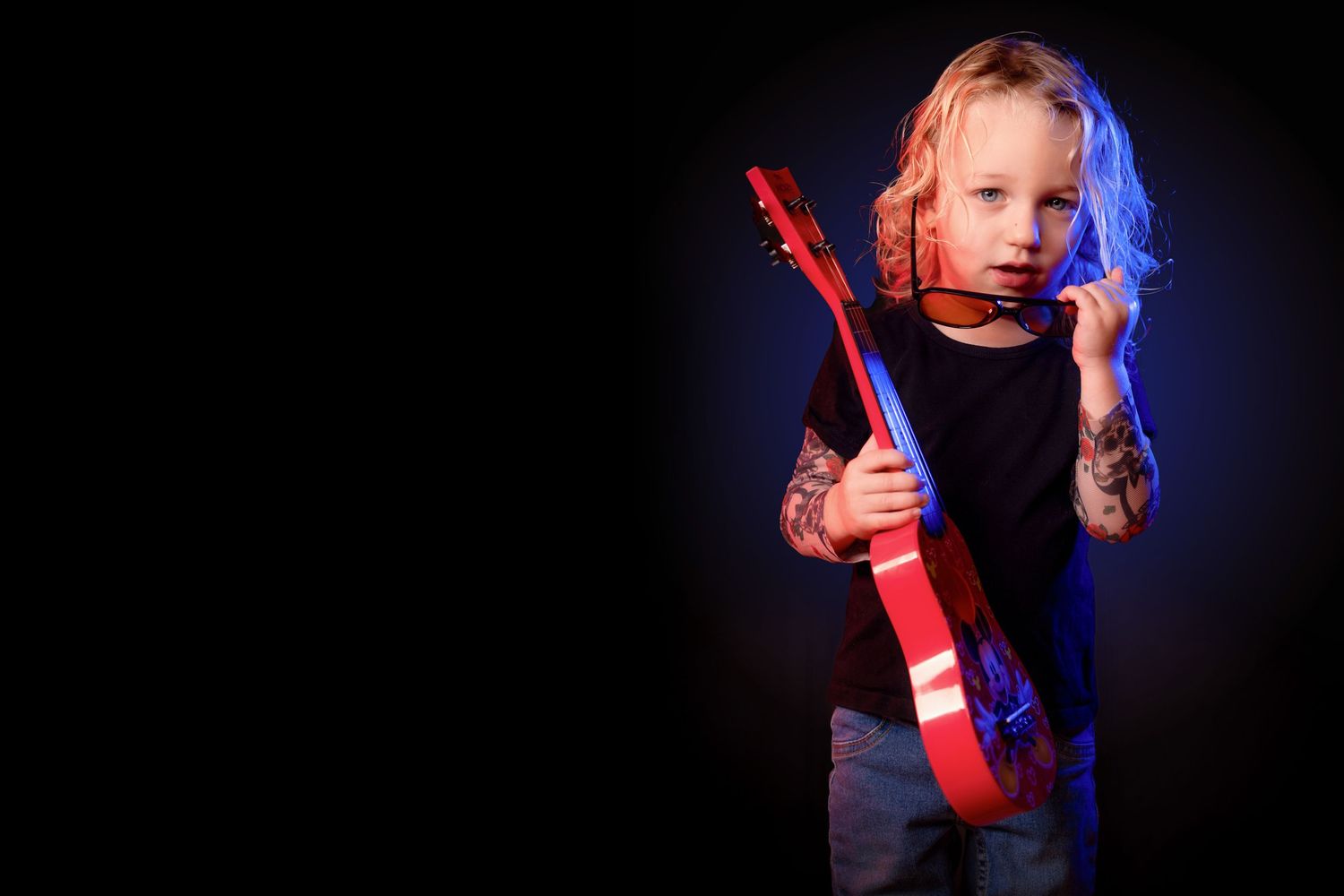 Cool blonde child with fake tattoos holding red electric guitar under blue stage lighting on black background.